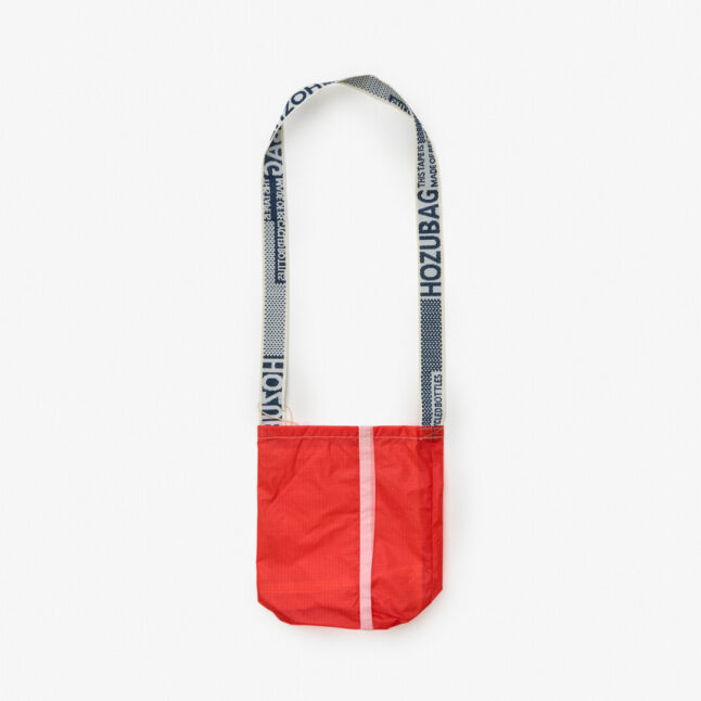 A red shoulder bag made of recycled paraglider fabric. The shoulder strap features a blue and white pattern and a writing that says 'HOZUBAG', as well as 'This tape is made of recycled bottles.'