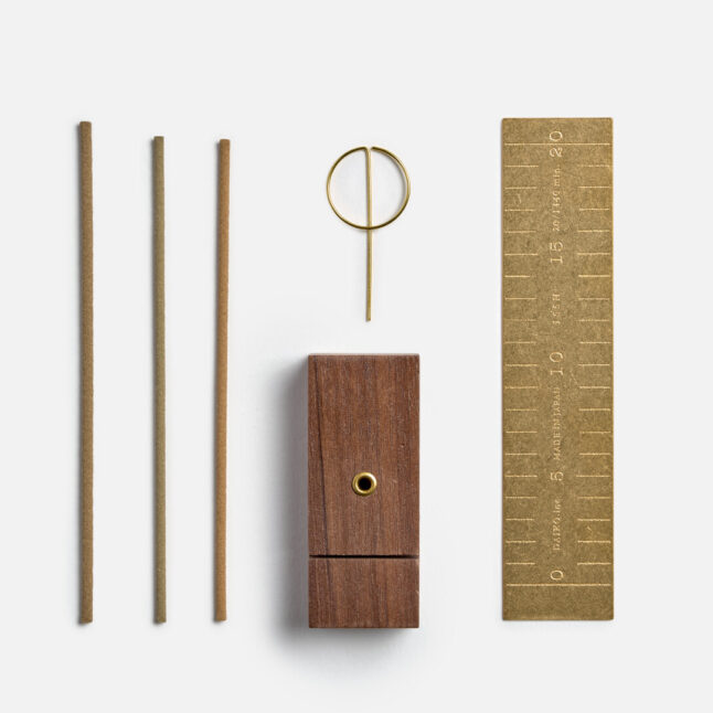 This image shows a minimalist incense set consisting of three incense sticks, a brass incense holder shaped like a loop, a wooden incense burner with a small hole, and a brass ruler marked with measurements from Japan House London. The items are arranged neatly against a clean, white background.