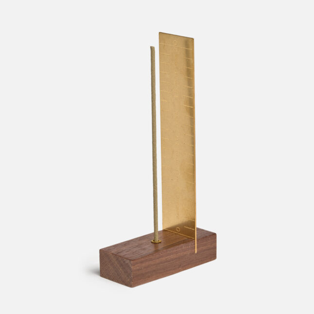 A brass measure with a wooden base holding a stick of incense from Japan House London.