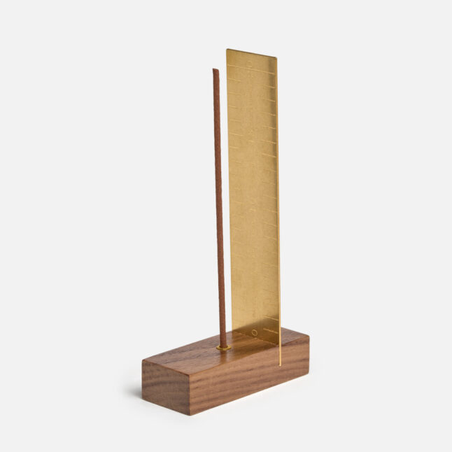 A brass measure with a wooden base holding a stick of incense from Japan House London.