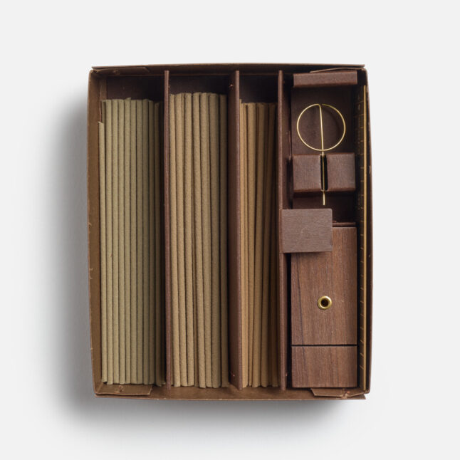 A boxed incense set neatly organized into compartments. It includes three bundles of incense sticks, a wooden incense burner, a brass incense holder, and a brass ruler from Japan House London. The natural tones of the materials give the set a simple yet elegant appearance.