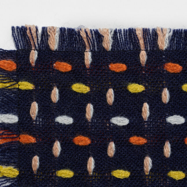 A wool table mat with a dark blue background and orange, yellow and white dots from Japan House London.