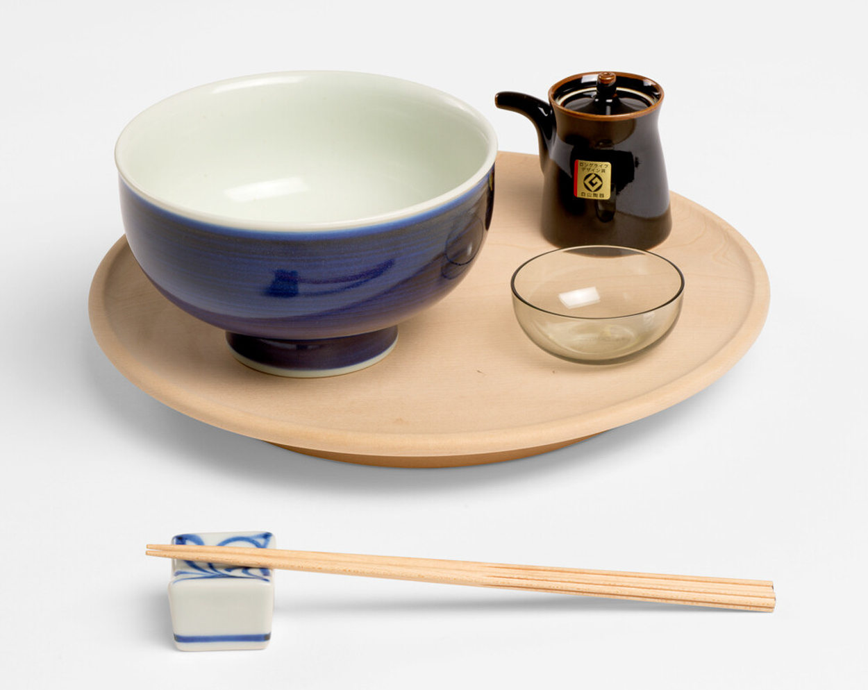 A large blue ceramic bowl with a brown soy sauce pot and small glass dipping bowl on top of a light wooden tray. There are a pair of light wooden chopsticks on a ceramic white and blue chopstick rest.