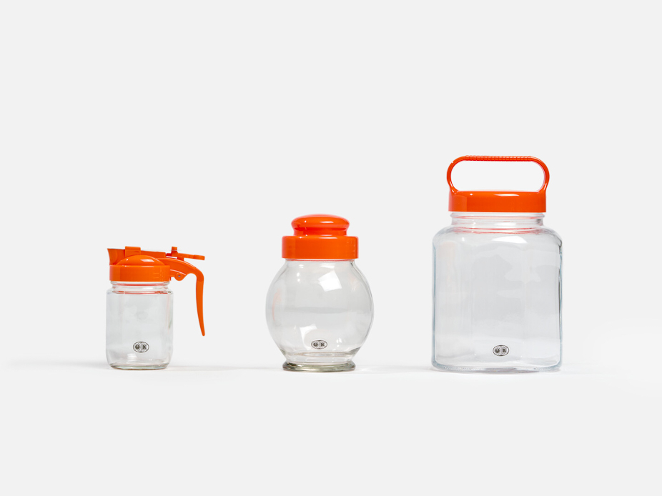 A glass server with an orange lid next to a rounded glass with an orange lid and the last glass container has an orange lid with a sandal