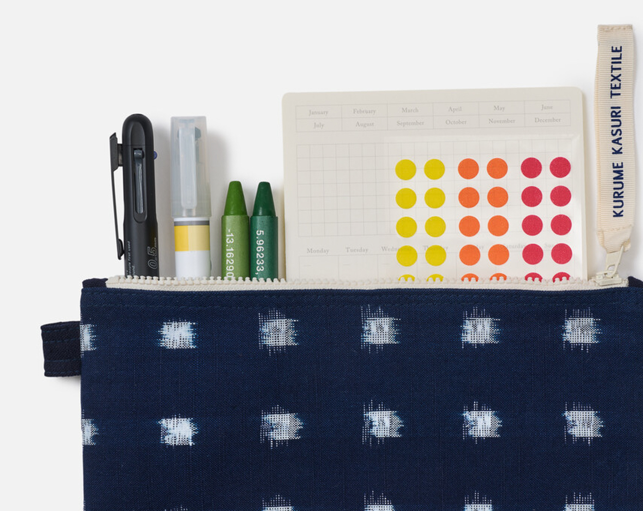 An indigo dyed pencil case with round stickers of red orange and yellow coming out along with two green crayons, one yellow highlighter and one green pen. The tag on the pencil case says Kurume Kasuri Textiles,