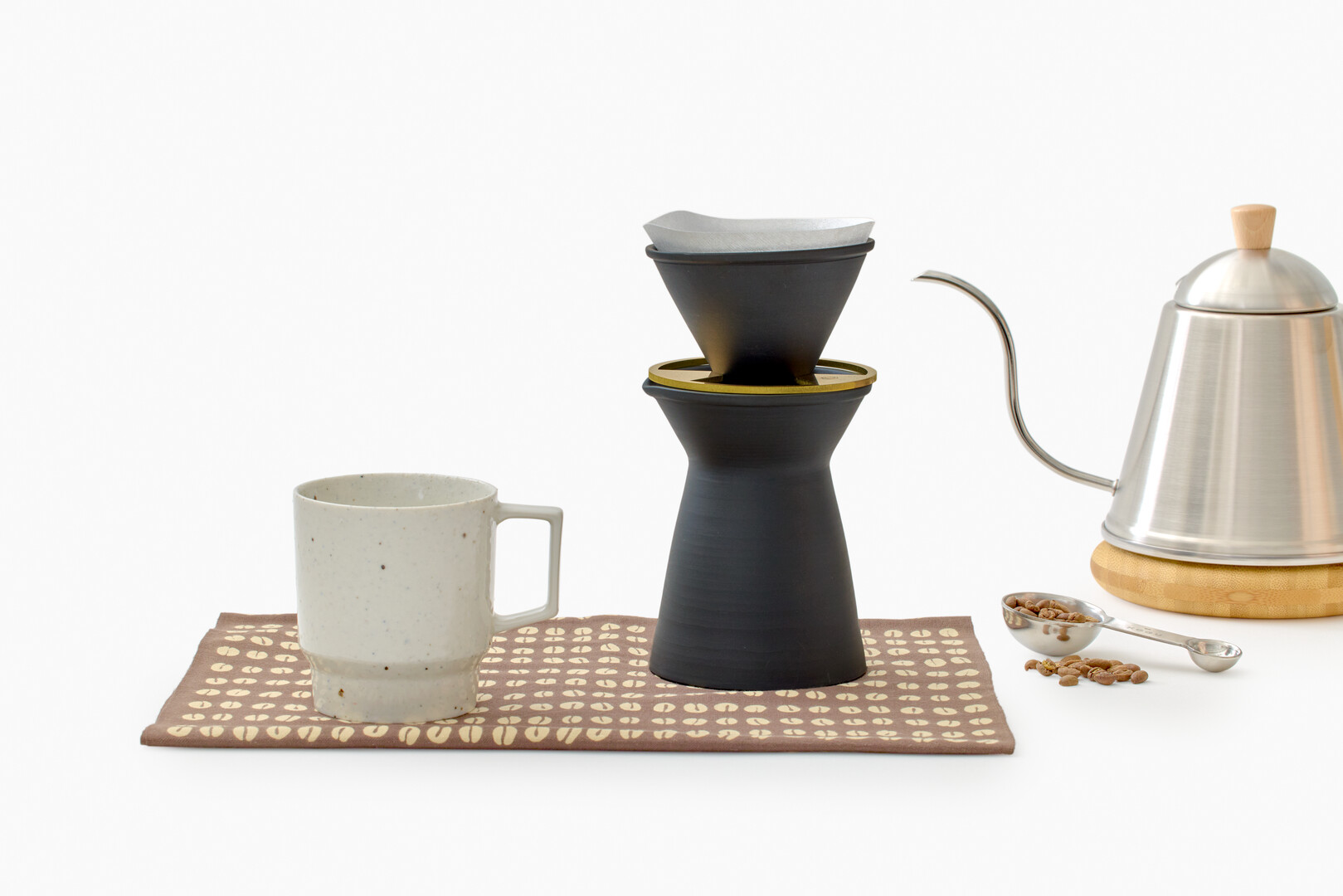 A white speckled mug with a black coffee carafe next to it with a metal silver kettle