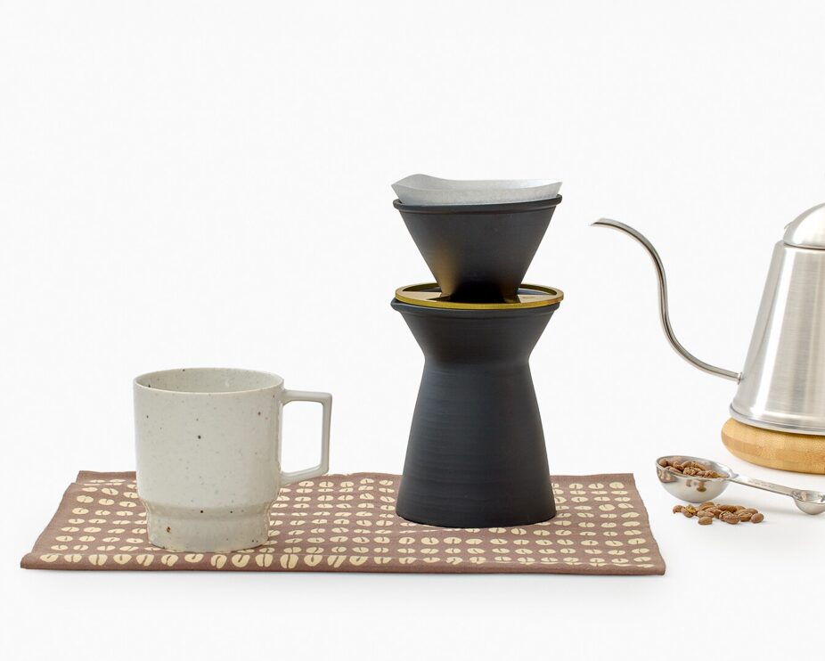 A white speckled mug and black coffee carafe sitting on a dark brown tenugui with a coffee bean pattern. Next to them is a pile of coffee beans with a coffee measure and stainless steel coffee pot with a thin spout.