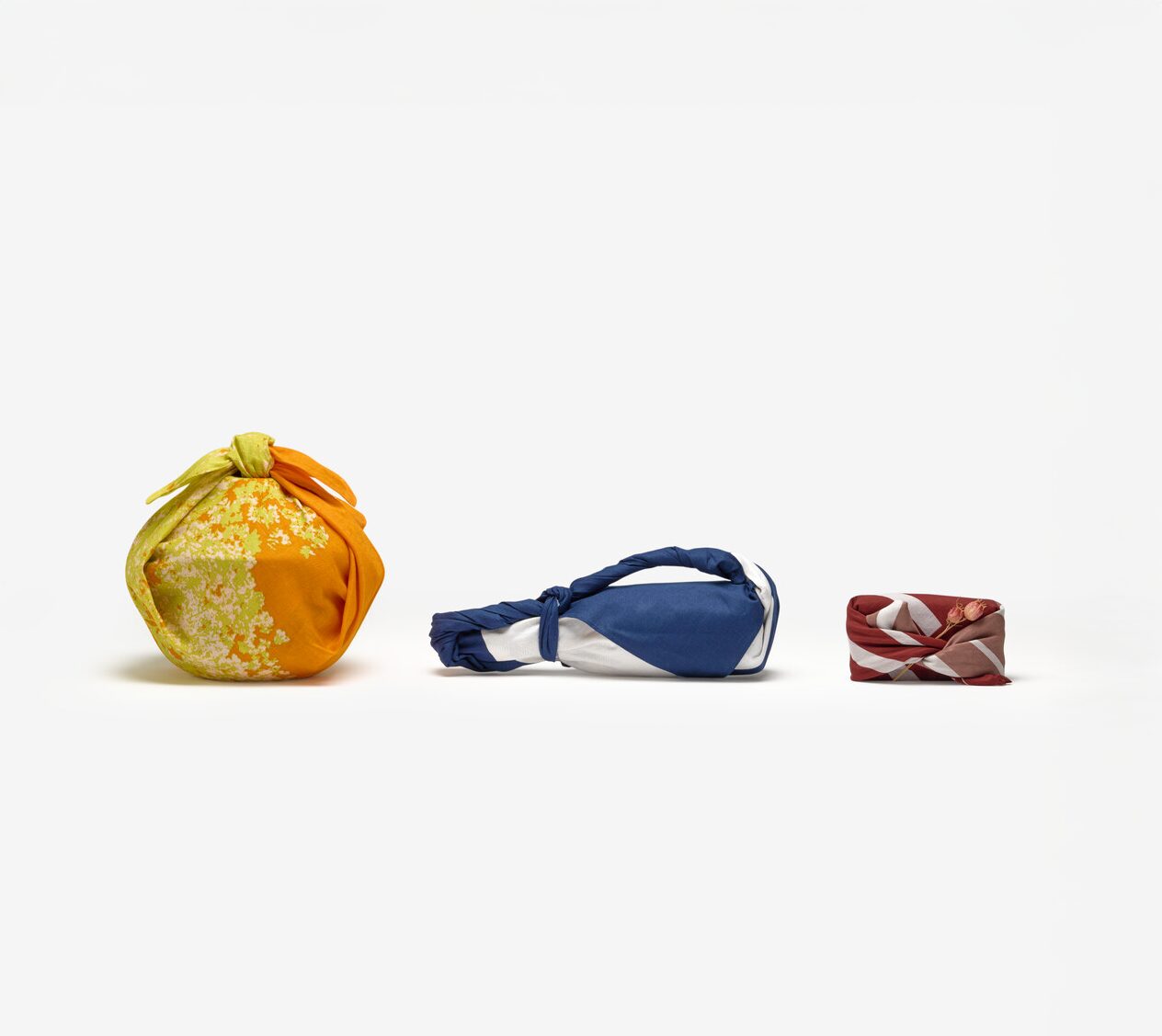 Three objects wrapped in a yellow and orange furoshiki, a navy and white furoshiki and a dark red and white furoshiki