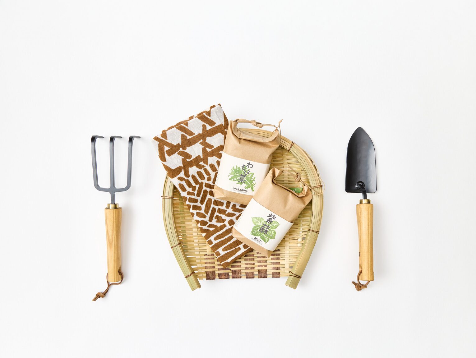 A hand fork for gardening with a basket holding a tenugui with a woven pattern and two paper bags of seeds and a hand shovel next to the basket