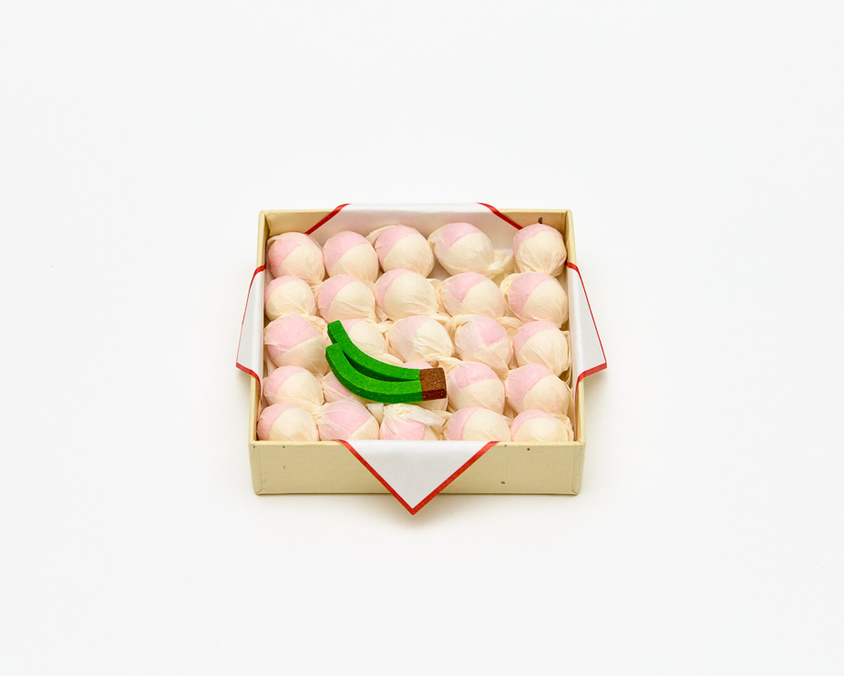 A small paper box with rows of small round sweets which are half pastel yellow and half pastel pink with two green stems on top