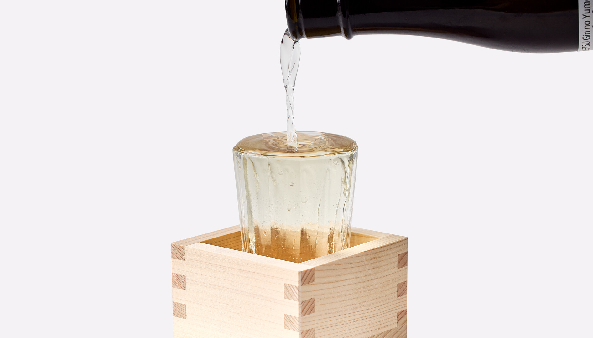 Sake being poured from a brown bottle into a small glass cup sitting inside a wooden cube