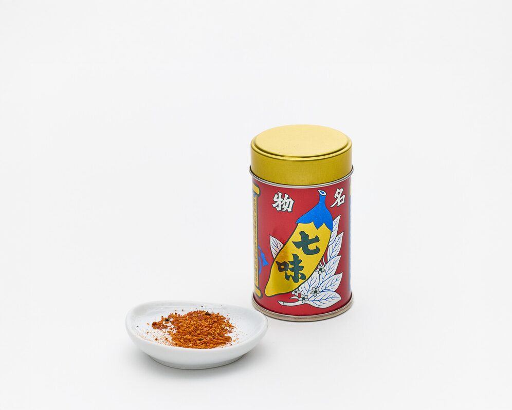 A red tin with a gold lid with shichimi in Japanese characters and a small plate with red ground spices