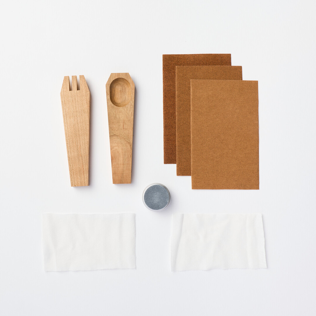 A piece of wood roughly shaped to be a fork next to a piece of wood roughly shaped as a spoon. They are next to three grades of sandpaper and above two white clothes and a small tin of wax