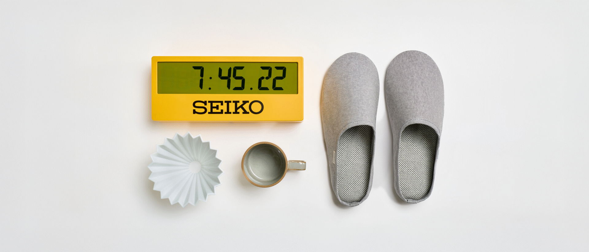 A yellow SEIKO mantle clock next to a grey pair of Sasawashi slippers and a white Origami ceramic coffee filter and ceramic grey Hasami mug with a beige edge.