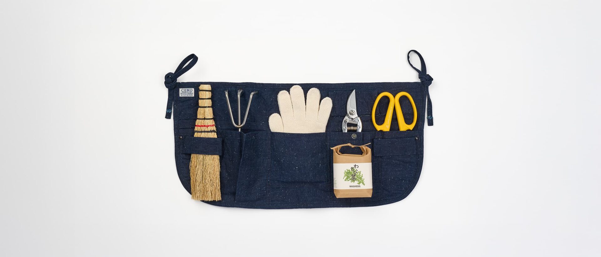 A short indigo gardening apron with pockets filled with a hand brush, a hand rake, a gardening gloves, a pair of shears and flower shears. Also with a packet of shungiku seeds on top.
