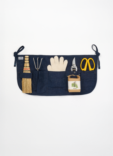 A short indigo gardening apron with pockets filled with a hand brush, a hand rake, a gardening gloves, a pair of shears and flower shears. Also with a packet of shungiku seeds on top.