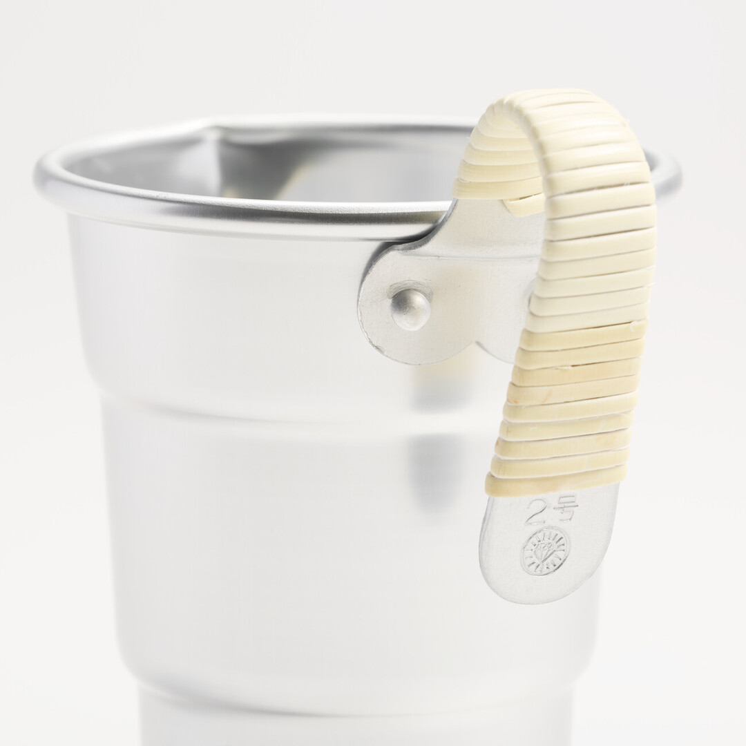 An aluminium sake pitcher with a bamboo wrapped handle. There is a detail of stamped at the end of the handle with the diamond mark of the brand and the words 2 gō