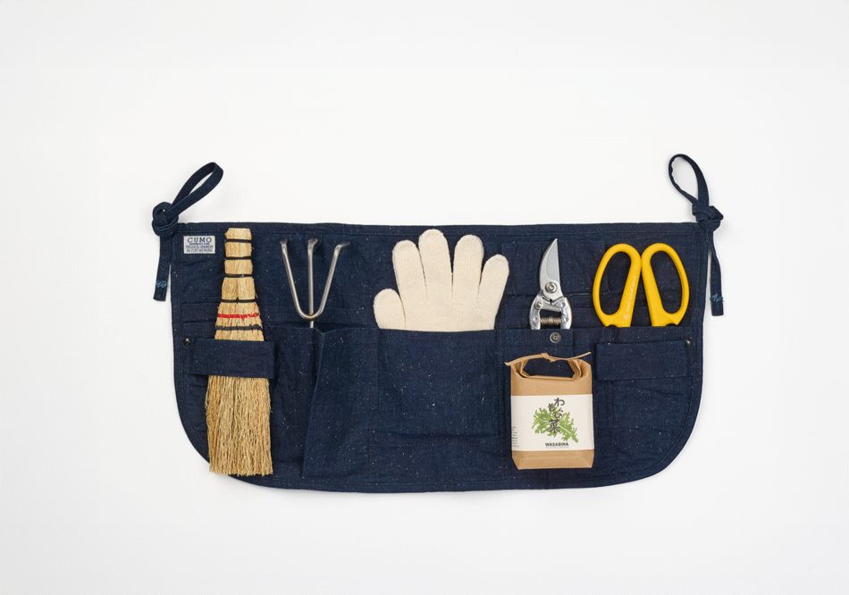 A short indigo gardening apron with pockets filled with a hand brush, a hand rake, a gardening gloves, a pair of shears and flower shears. Also with a packet of shungiku seeds on top.