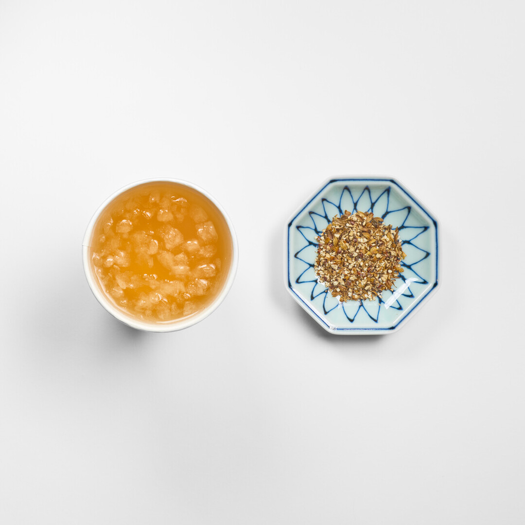 A amber coloured barley tea in a cup with ice and next to it is a octagonal plate with barley grains