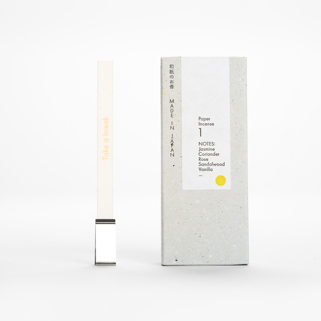 A think rectangular washi paper incense in No. 1 - Jasmine, Coriander, Vanilla, clipped in a silver clip next to a grey box from Japan House London.