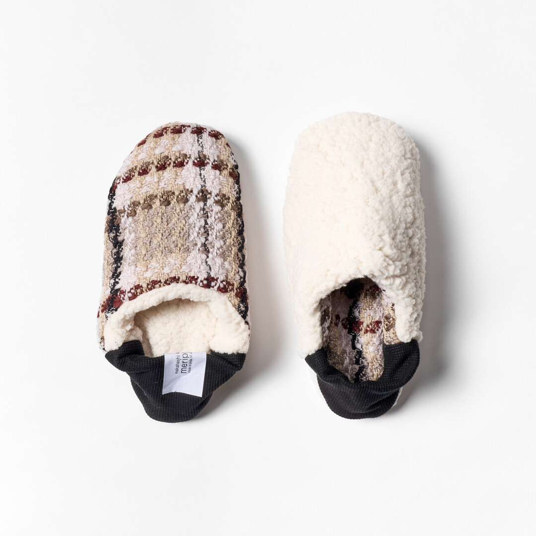 A pair of reversible slippers with a beige and brown tartan fabric and white sheep boa fabric on the other side with a black ribbed heel from Japan House London.