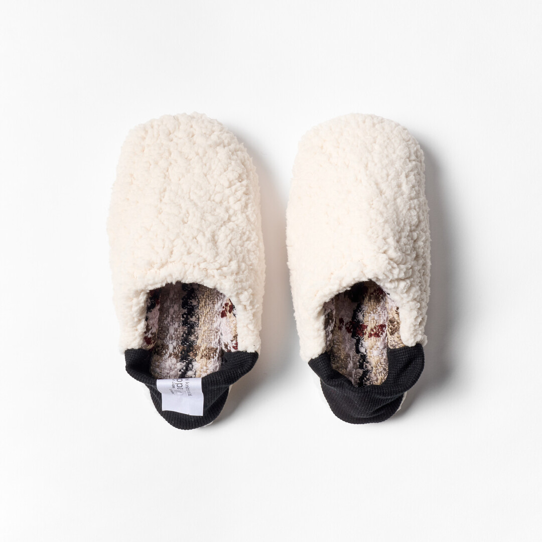 A pair of reversible slippers with a beige and brown tartan fabric and white sheep boa fabric on the other side with a black ribbed heel from Japan House London.