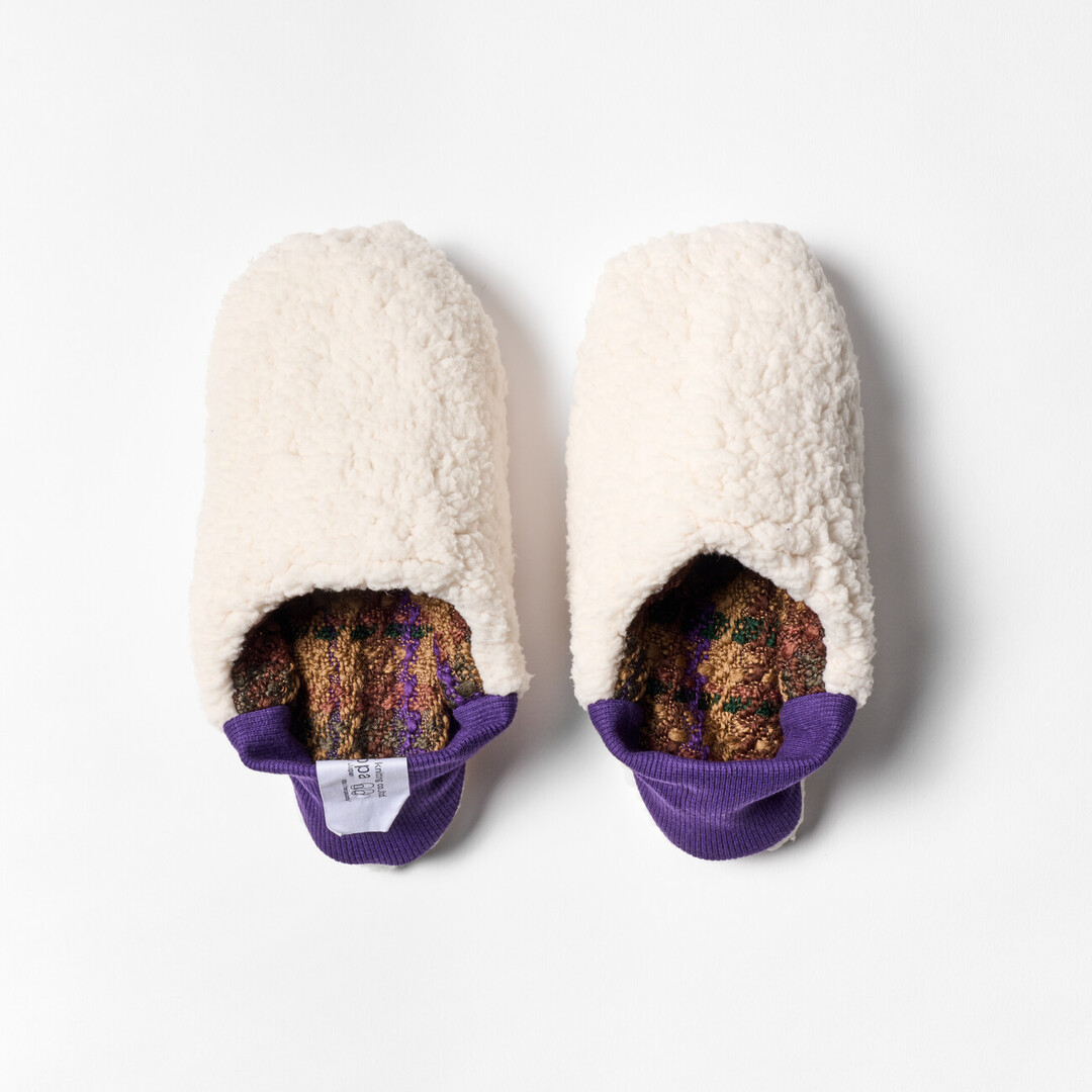 A pair of reversible slippers with a green and brown tartan fabric and white sheep boa fabric on the other side with a purple ribbed heel from Japan House London.