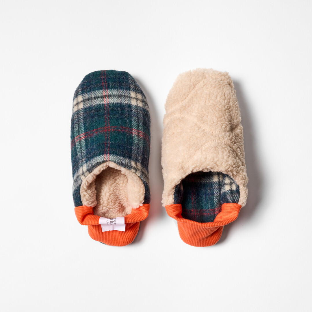 A pair of reversible slippers with a blue and green tartan fabric and beige shaggy fabric on the other side with a orange ribbed heel from Japan House London.