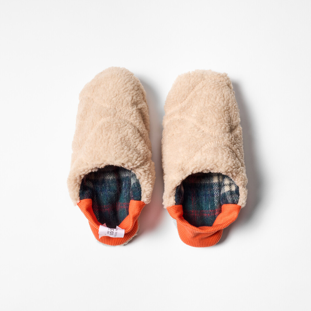 A pair of reversible slippers with a blue and green tartan fabric and beige shaggy fabric on the other side with a orange ribbed heel from Japan House London.