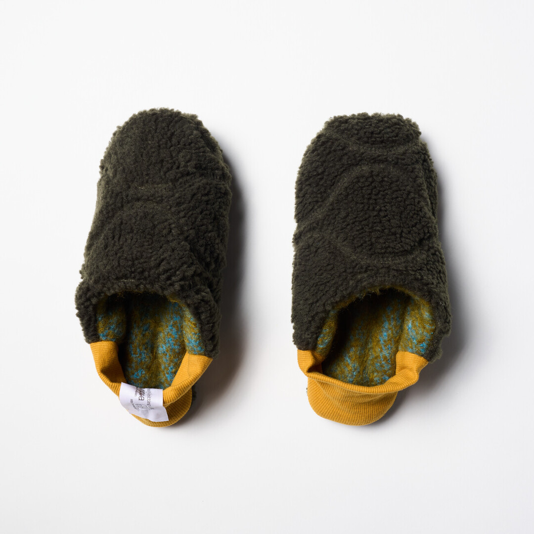 A pair of reversible slippers with a felted green and blue blended fabric and a dark green shaggy fabric on the other side with a yellow ribbed heel from Japan House London.