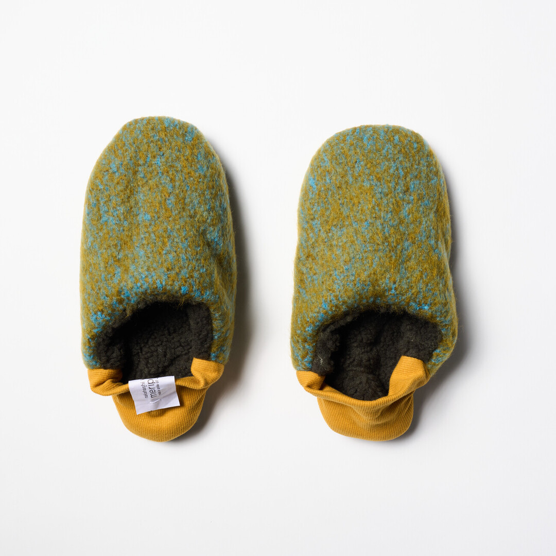 A pair of reversible slippers with a felted green and blue blended fabric and a dark green shaggy fabric on the other side with a yellow ribbed heel from Japan House London.