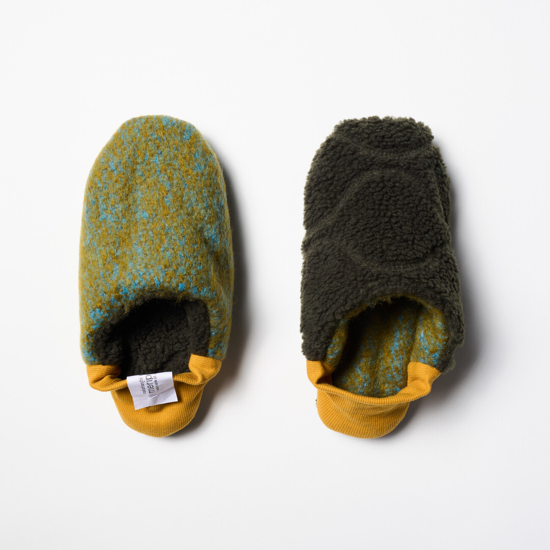 A pair of reversible slippers with a felted green and blue blended fabric and a dark green shaggy fabric on the other side with a yellow ribbed heel from Japan House London.