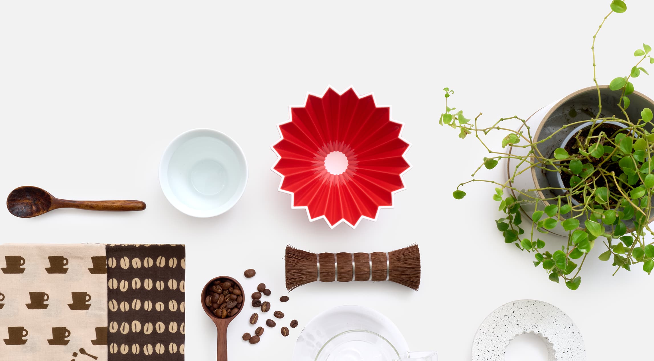 Coffee products of a red ceramic origami coffee dripper, a natural fiber brush, a wooden coffee spoon, two brown coffee themed tenugui and some coffee beans scattered from Japan House London.