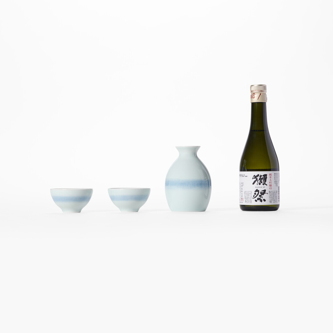 A small bottle of Dassai with two white with a blue detail along the middle sake cups and a sake serving vessel with the same patterns as the cups from Japan House London.