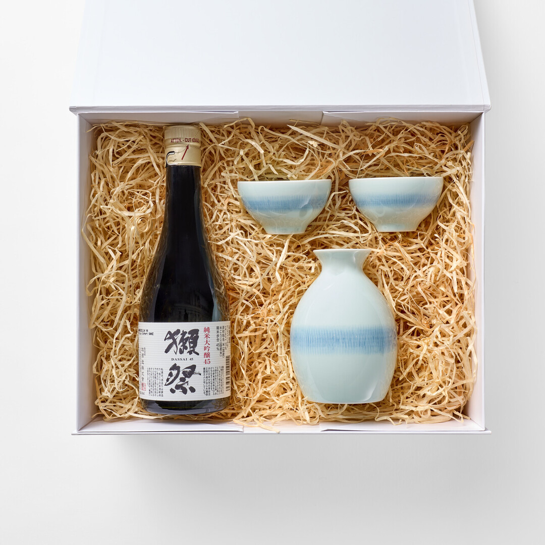 A white gift box with a small bottle of Dassai with two white with a blue detail along the middle sake cups and a sake serving vessel with the same patterns as the cups in wood shavings from Japan House London.