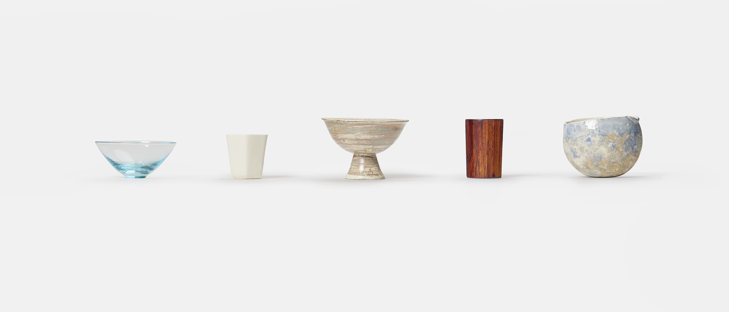 Five small vessels made from different materials of ceramics, wood and glass in a row from the exhibition Hyakko 100 Makers from Japan House London.