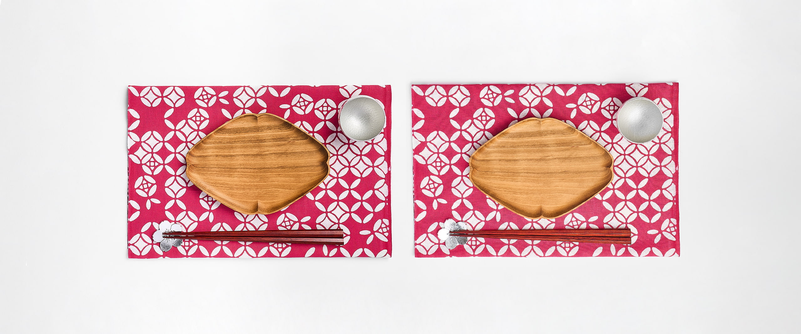 Two sets of diamond shaped wooden plate alongside chopsticks with chopstick rest and a tin sake cup under a patterned cloth from Japan House London.