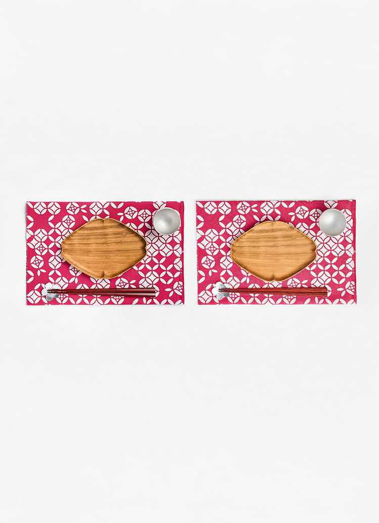 Two sets horizontally next to each other of a diamond shaped wooden plate, chopsticks with tin chopstick rests and tin sake cups on a pink patterned material from Japan House London.