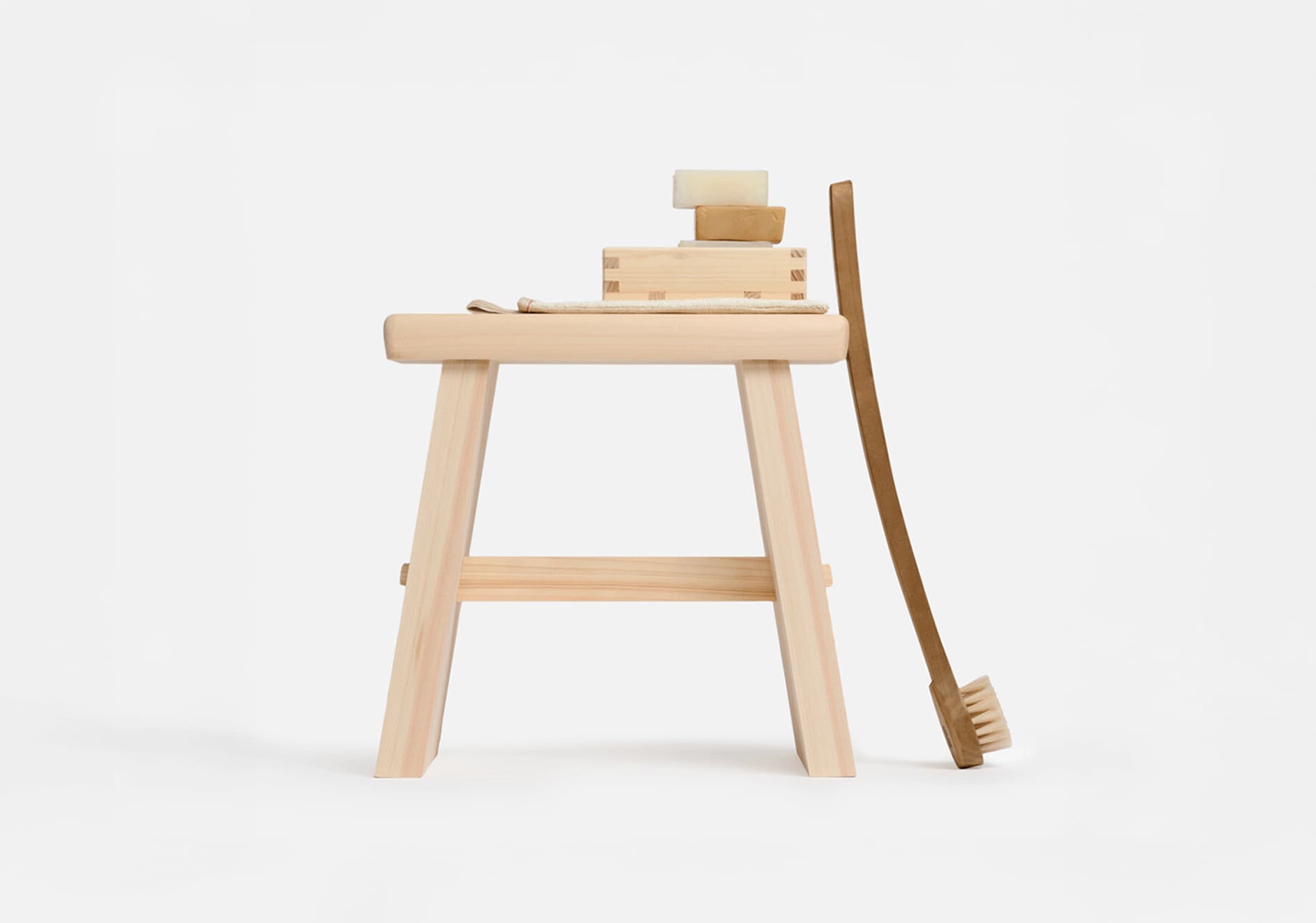 A wooden bath stool with a wooden soap holder on top with a stack of soap with a body brush leaning against the stool from Japan House London.
