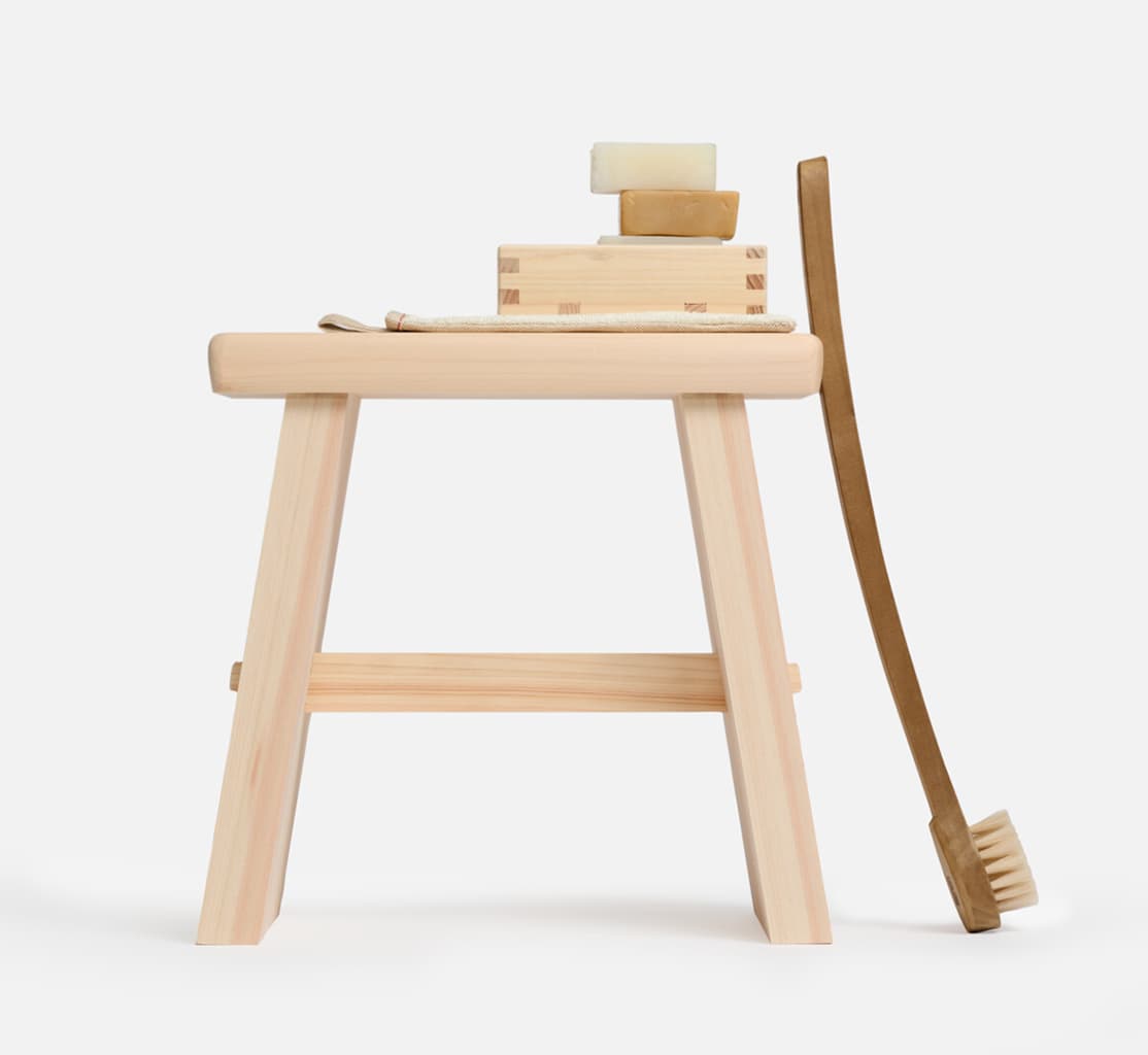 A wooden bath stool with a wooden soap holder on top with a stack of soap with a body brush leaning against the stool from Japan House London.