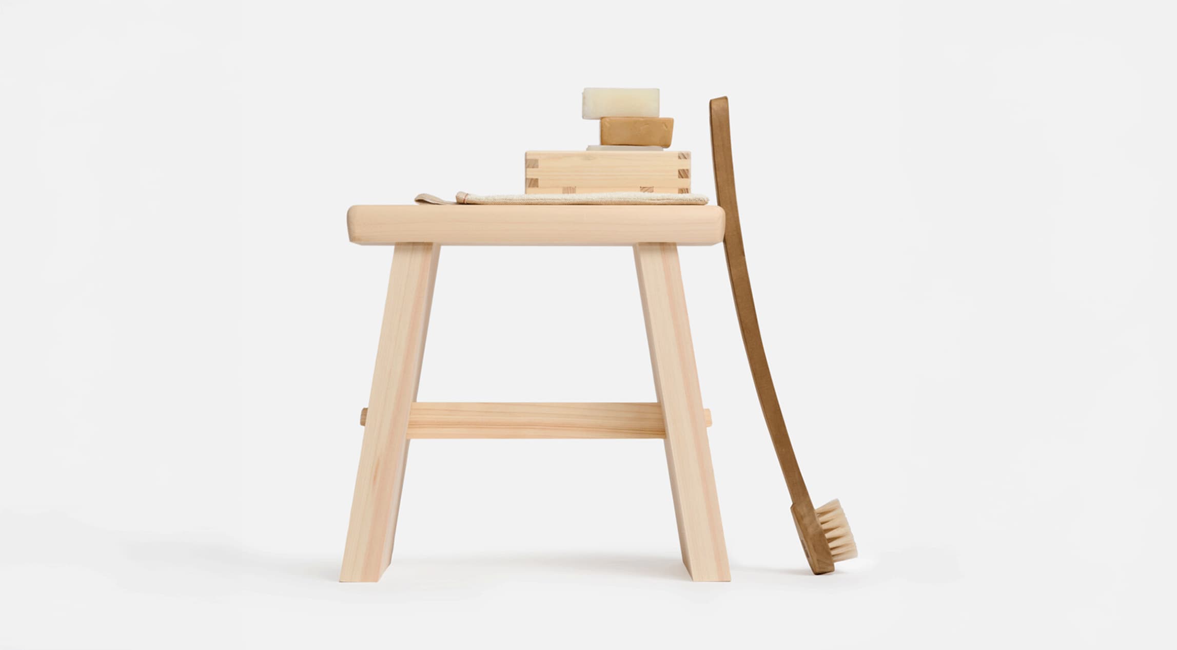 A wooden bath stool with a wooden soap holder on top with a stack of soap with a body brush leaning against the stool from Japan House London.