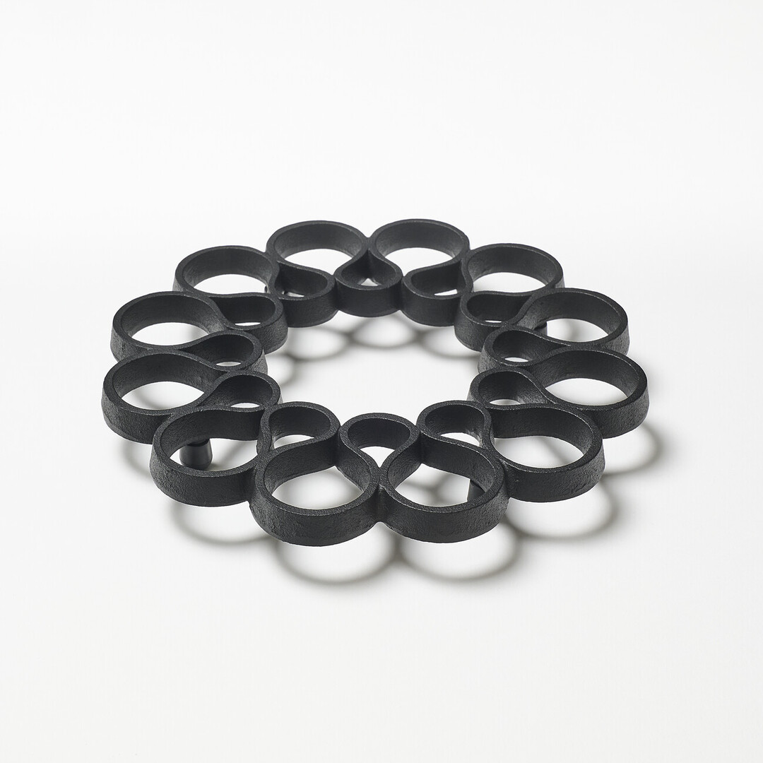 A black iron trivet shaped with undulating lines in a donut form by OIGEN Foundry Co., Ltd. from Japan House London.