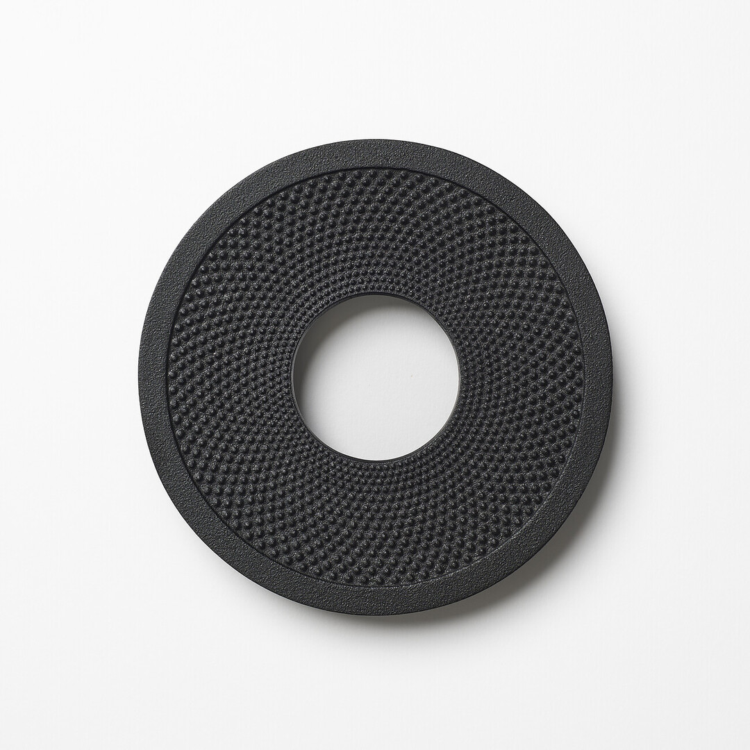 A cast iron black trivet shaped like a donut with a textured surface by OIGEN Foundry Co., Ltd. from Japan House London.